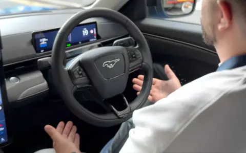 A man is sitting in the driving seat of a car but his hands are off the wheel.