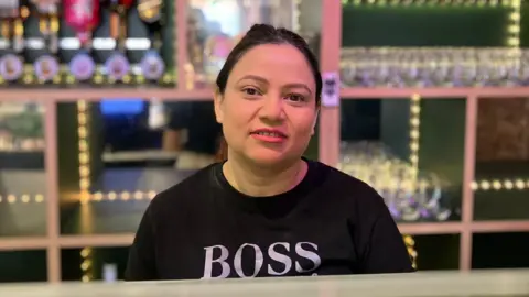 A woman in front of a bar with optics behind her. She wears a black T-shirt with a BOSS logo.