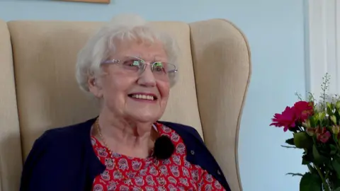 A picture of Beryl sat in a large cream coloured chair. She is wearing a red outfit with a navy blue cardigan. She is wearing glasses, while smiling.