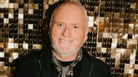 Huw Rees Huw Rees is smiling, wearing a dark jacket and top with grey and brown patterned silky cravat. He has grey hair and a beard. He is standing against a gold tiled backdrop.