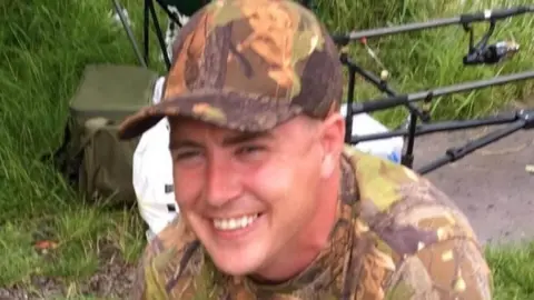 Undated handout photo of Paul Smith, whose body was found in a wheelie bin in Everton. He is wearing camouflage patterned baseball cap and t-shirt with fishing equipment in the background. He is smiling. 