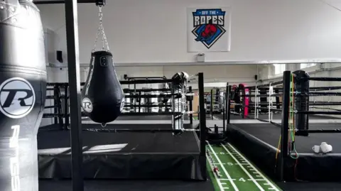 A gym with two boxing rings, a green lane in the middle for weight pushing, and punching bags. The background features wall mirrors.