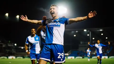 Will Cooper/Bristol Rovers FC Jamie Lindsay smiles at fans with his arms outstretched as he scores the only goal of Bristol Rovers' 1-0 win over Shrewsbury at the Memorial Stadium. Other Rovers players also run to join the celebrations.