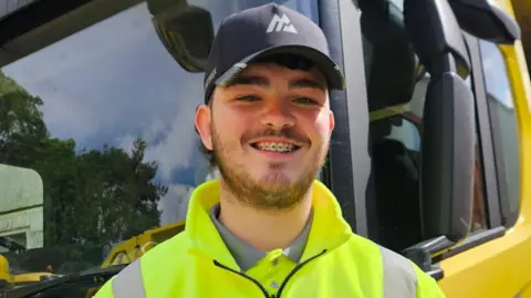 Gaskells Daniel Kinsella has a dark beard and is wearing a black baseball cap and yellow high-vis jacket. He is standing in front of a yellow bin lorry and smiling for the camera.