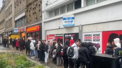 Simon Thake/BBC A large queue of people wrapped up in warm clothes  stand on a grey day outside a series of shops