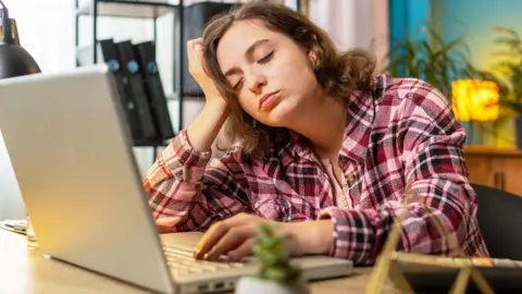 A woman with a brown bob, wearing a pink flannel shirt, sits at a table in front of an open laptop. She is resting on hand on the keyboard and the other is propping her head up. Her eyes are closed.