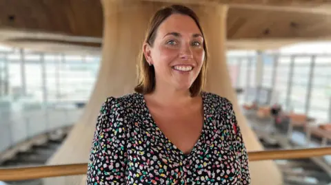 The image shows Heledd Fychan standing in the Senedd looking directly at the camera and smiling. She is wearing a colourful dress. The Senedd's funnel is behind her.