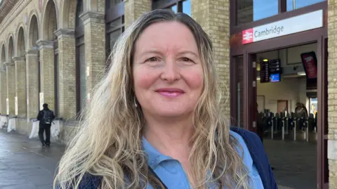 Tom Jackson/BBC Sallie Bates is standing in the middle. She is outside of Cambridge's train station and has long blonde hair. She is wearing a blue jacket.