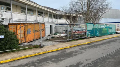A building site in front of Guernsey's Princess Elizabeth Hospital. On the site there are a number of shipping containers and metal poles.
