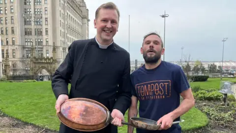  Rector of Liverpool Father Philip Anderson is stood smiling at the camera with a copper frying pan as he presents it to Lee Johnson