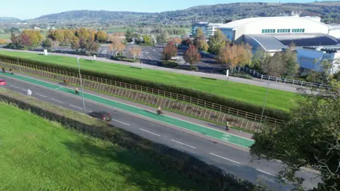 A computer generated image of cyclists using a cycleway alongside a main road in the Gloucestershire countryside. Cheltenham Racecourse and Cleeve Hill can be seen in the background on what is a sunny day.