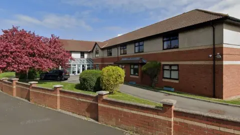 Google A Google Street View screenshot of Silver Mews nursing home in Redcar. The two-storey building has red bricks on the lower ground and rendered cream/grey walls on the upper floor. There is a small patch of glass with a tree and some shrubs in front on the property. 