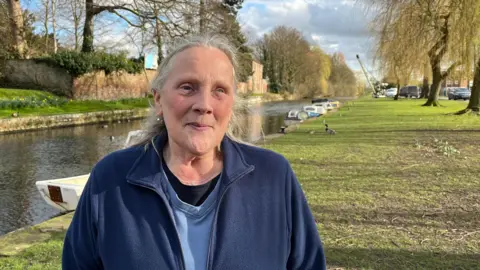 A woman stands beside a canal. She has long grey hair and is wearing a dark blue fleece jacket. She is standing on a grass bank lined with large willow trees. The canal is in the background with several boats moored alongside it.