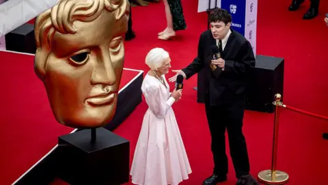 Getty Dame Mary Berry holds a microphone in one hand as she speaks to Baftas red carpet host Roman Kemp. Roman is wearing a black suit with a white shirt.