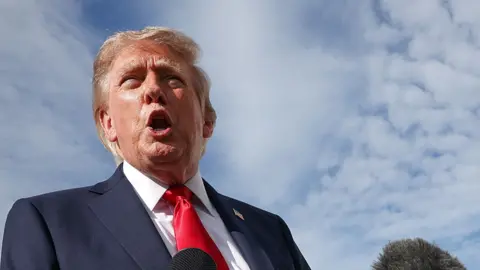 US President Donald Trump, a blond man wearing a navy suit with a red tie, speaks into a microphone.
