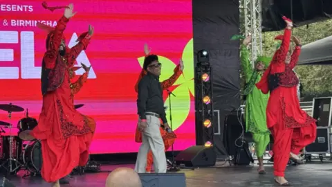 A group of performers on stage. A man in a black shirt and cream trousers stands in the middle, surrounded by dancers in colourful clothes.