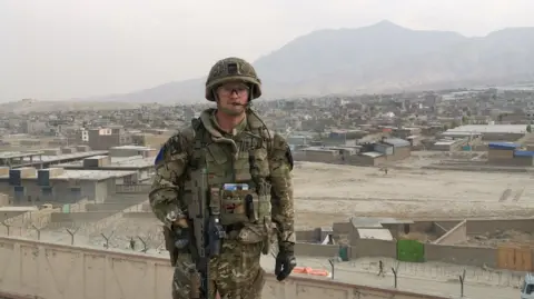 Aaron Stone A soldier wearing a camoflauge army suit and holding a rifle. He is stood in front of a dust covered compound, with a mountain in the background.