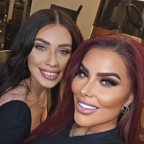 Leanne Baikie Two women with styled hair and full makeup smile at the camera while taking a close-up selfie inside a salon. One has long dark hair, and the other has long red hair and visible arm tattoos. Salon chairs, mirrors, and styling tools can be seen in the background.