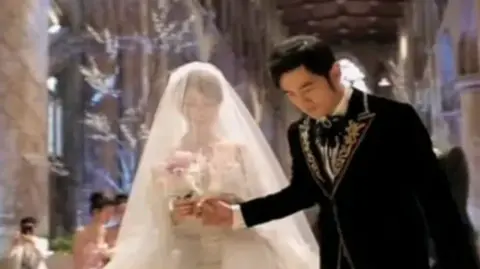 Other A woman wearing a wedding dress with a veil over her head stood next to a man wearing a black suit with gold detail. The woman is holding flowers as well as holding the man's hand. They are both stood in a church setting. 