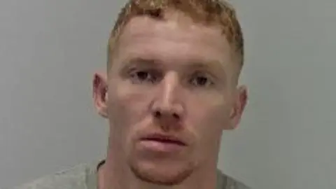 A man with ginger hair and a small ginger beard against a grey background