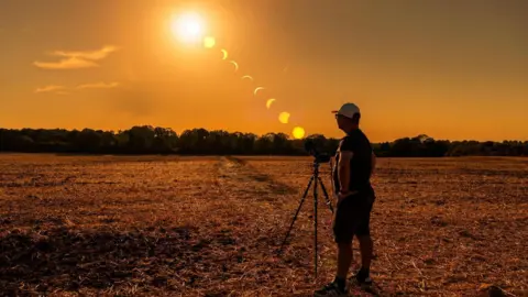A person stands beside a camera on a tripod in a harvested field in Caterham, Surrey, during sunset. A sequence of the eclipse is shown across the sky, tracking the Sun as the Moon moves across it from high above the horizon to near sunset. Trees line the horizon beneath an orange sky.