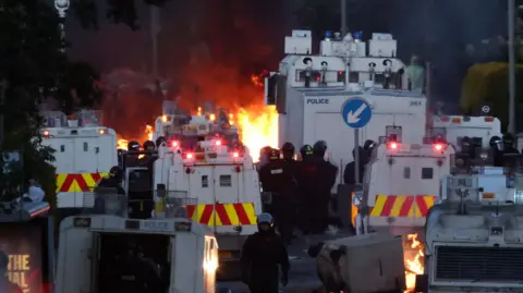 Police officers wearing riot gear stand beside vans as fires are lit in the distance.