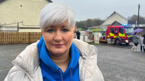 Joanne Bunting wearing a white puffer coat and blue hoodie she has short white hair. Behind her is a fire engine