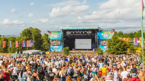 A large crowd begins to gather in front of the main stage in a field in the sunshine. There are colourful flags around the stage.