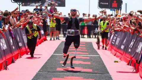 Getty Images A male double amputee crosses the finish line of a race with his arms stretched wide as people cheer and applaud. 