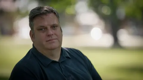 Steven is wearing a blue shirt and is sitting on a bench outside. He is in focus but the background, which resembles grass and trees, is fully out of focus. Steven is looking directly into the camera with a straight face. 