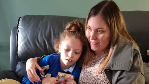 Dave Clarke Addy being hugged by her mother on a leather sofa. Addy wears a blue jumper and is holding what looks like a wrapped present, while her mother Hayley wears a khaki jacket. 