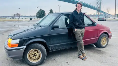 A man is in a car park leaning on a car made of two front ends - one red and one black. A bridge is in the background. 