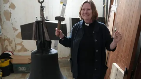 A woman vicar in black, wearing her dog collar, smiles at the camera. She has brown hair in a bob. She is holding a mallet in one hand and is standing next to a church bell 