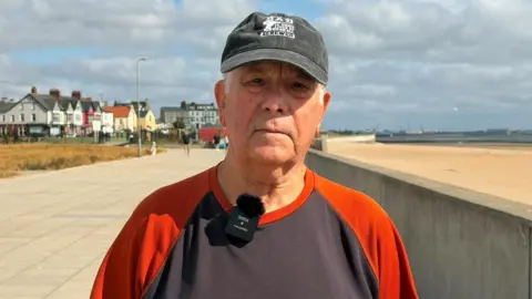 Adrian Barron wears a black cap and a black t-shirt with orange sleeves. He is standing on the promenade next to the sandy beach. 