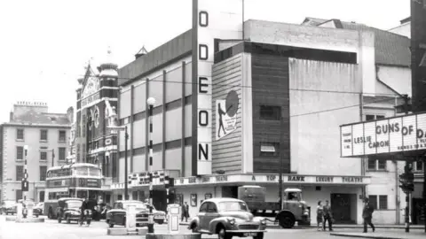 Northern Ireland Historical Photographical Society A black and white image of a busy street with old fashioned cars and a concrete theatre with a ODEON sign at the front