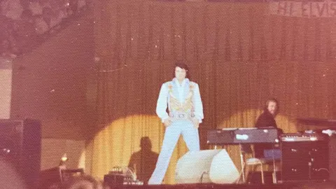 Jan Owen Elvis Presley in concert in 1976. The photo, by Jan Owen, shows Presley dressed in a light-colored jumpsuit with gold embellishments, standing on a stage. The background features a large curtain, and there are musicians and equipment, such as keyboards, on stage to the right. 