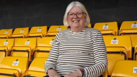 A woman with light blonde hair and a white and black striped top smiles as she sits on a yellow chair in a football stand.