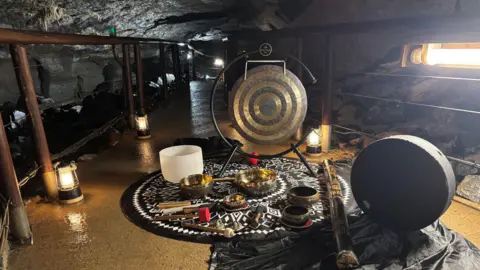 BBC A mixture of large and small gongs, metal bowls and wooden rain sticks are placed on a circular mat. In the background is the Marble Arch Caves