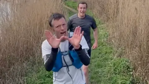 Max Howard The picture shows two people running along a narrow, grassy path surrounded by tall, golden reeds. The person in front is holding both hands up in front of the camera, as if gesturing or playfully shielding the view. This runner is wearing a white t-shirt, with dark long-sleeved top underneath, blue shorts and a hydration vest.