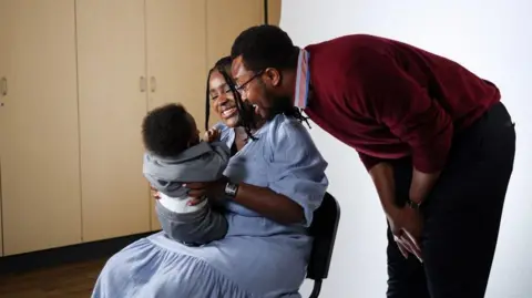 Redcar and Cleveland Council/Middlesbrough Council Lorna Mulungi with her husband, Yassir, and their baby. Lorna is seated and has long black hair. She is wearing a light blue dress and holding the child. Yassir has short dark hair and is wearing a dark red jumper and black trousers. He is leaning in as the pair smile towards their baby.