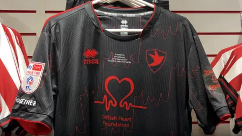 A photo showing a black football shirt with a red British Heart Foundation logo on the front. The shirt is hanging on a row in a shop surrounded by other football shirts