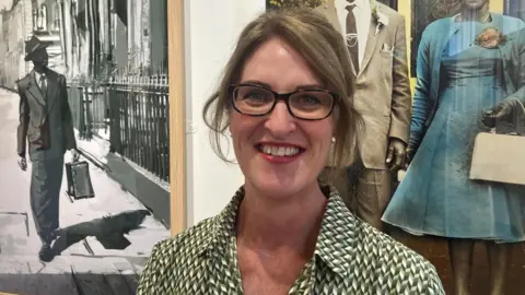 A woman wearing glasses standing in an art gallery, with framed photographs and artwork displayed behind her.