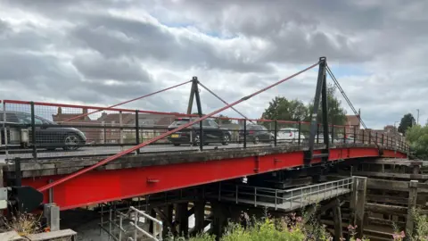A bridge over The River Ouse leading into the town of Selby. The bridge's metal base, which supports the road, is painted red. The railings are black and there are large 'V' shaped metal supports on each side of the bridge. Traffic is heading in both directions.
