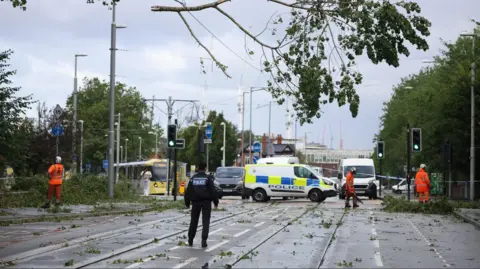 Reuters A police officer looks on as workers begin to remove fallen tree branches after strong winds brought by Storm Lilian brought down trees blocking roads and tram routes in Manchester.