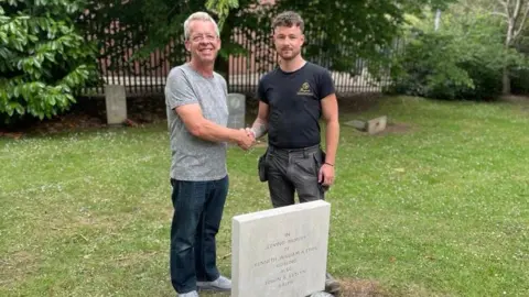A man with short grey hair wearing a grey patterned t-shirt and blue jeans stood next to a gravestone in a graveyard. He is shaking hands with a man with short brown hair who is wearing a black t-shirt and black trousers. The grass is green and green trees can be seen in the background. 