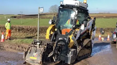 Wiltshire Council A large yellow machine being driven by a man in flourescent clothing on a muddy road.