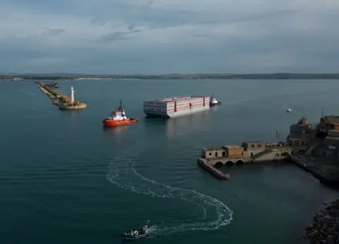 Dan Kitwood/Getty Images The Bibby Stockholm migrant barge (C) arrives at Portland Harbour on July 18, 2023 in Portland, England.