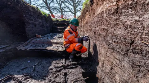 Wessex Archaeology A male archaeologist carrying out an excavation. He is wearing an orange high vis suit and a hard hat. He is kneeling down looking at the site.