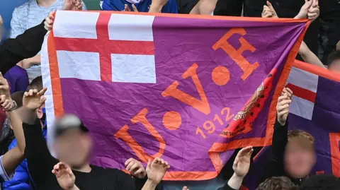 Getty Images A zoomed in image of a football crowd. Two peoples faces are blurred out and behind them a large purple flag with orange letters saying UVF are on the front. 