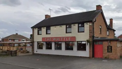 Google A red brick two-storey building with white and cream panelling on the front, as well as two rows of windows and a red sign reading 'The Whittle'. There is concrete in front of it, an outhouse on the right side and a seating area with wooden picnic benches, a fence and a canopy over the top.